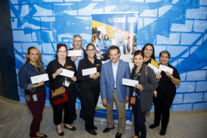 A group of six women and one man from Ensor Middle School hold up envelopes containing their gift cards next to CREEED Executive Director Eddie Rodriguez. They are all standing in front of a pop up banner with the CREEED logo